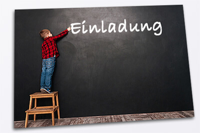 Postkarte auf der ein Junge zu sehen ist, der auf einem Hocker vor einer schwarzen Wand steht und Einladung mit Kreide an die Wand geschrieben hat.