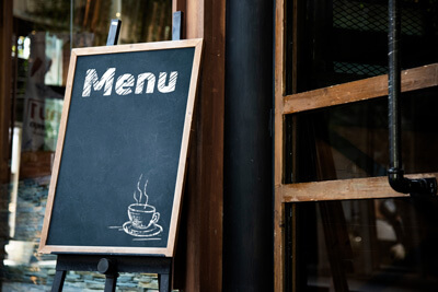 Eine Staffelei auf der eine Tafel steht vor einem Fenster. Auf der Tafel steht oben Menu und unten rechts ist eine dampfende Kaffeetasse auf der Tafel abgebildet.