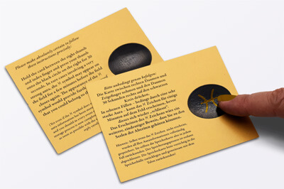 Zwei gleiche orangene Karten mit schwarzem Text. Rechts auf der Karte ist je ein schwarzes oval. Auf der einen Karte vollflächig, auf der anderen scheint das Logo durch.