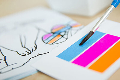 Die obere linke Ecke des Bildes ist verschwommen, auf dem unteren Teil des Bildes sieht man ein Stück von einem gezeichneten Osterhasen und einen Pinsel mit einer Wassermalfarbenkarte mit den Farben blau, pink und orange